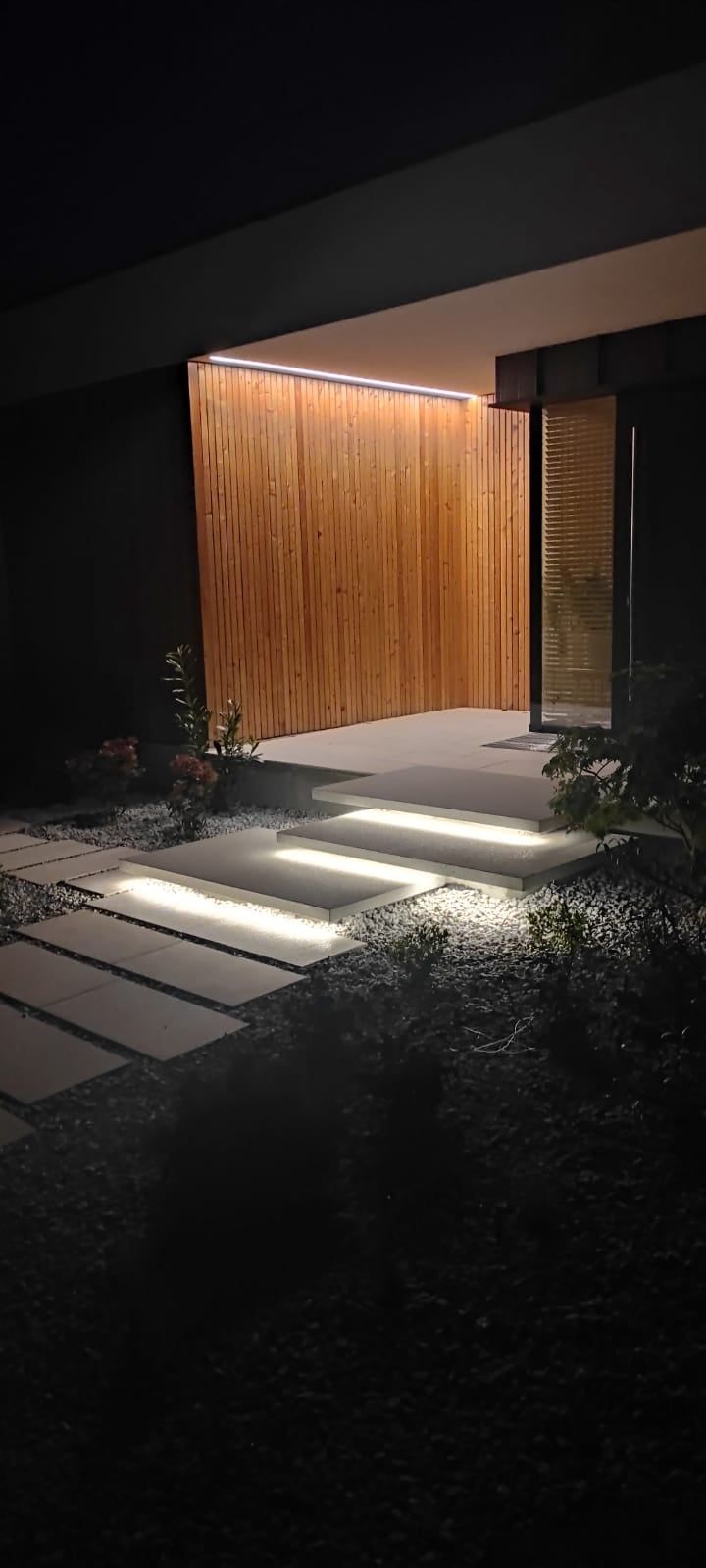 Wejście do domu nocą: taśmy LED pod stopniami i linia światła nad drewnianą okładziną